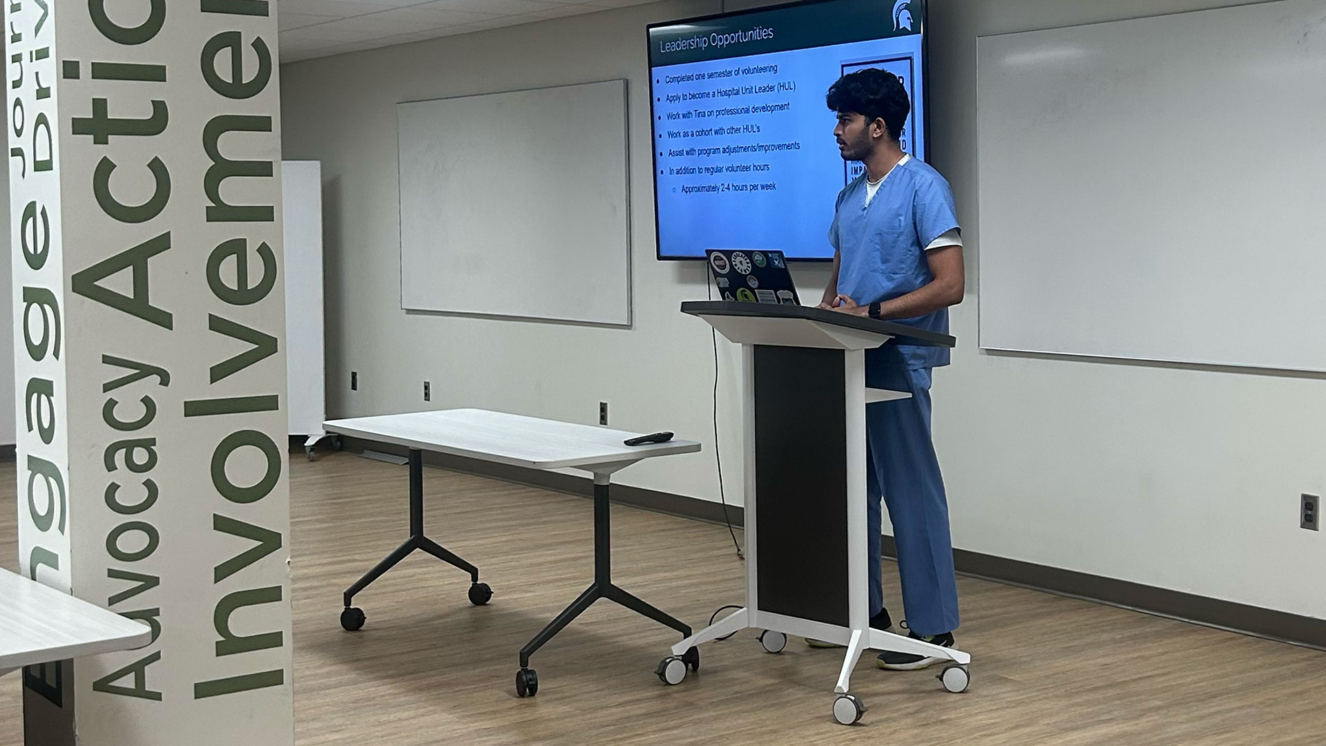 Student coordinator Karthick Prem Satheesh Kumar, wearing scrubs, leads an on-campus orientation for volunteers preparing to serve at McLaren Greater Lansing